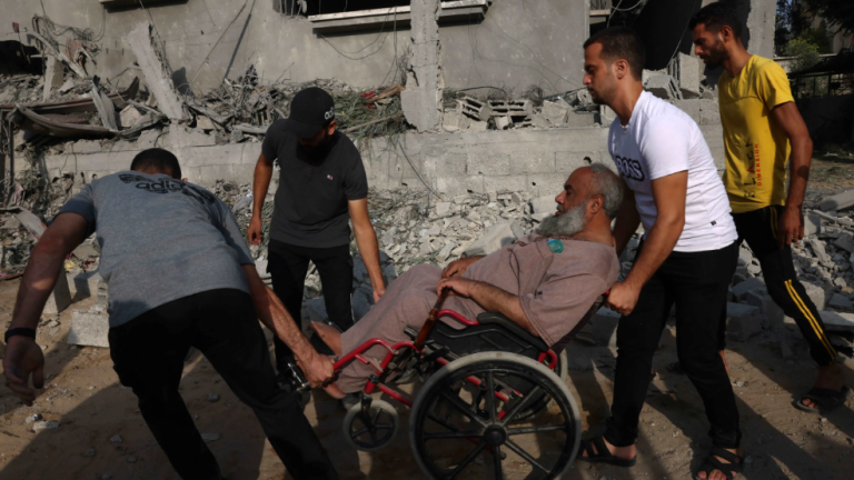 Tres hombres ayudan a un hombre en silla de ruedas a moverse entre los escombros y la destrucción en el sur de la Franja de Gaza el 22 de octubre de 2023.