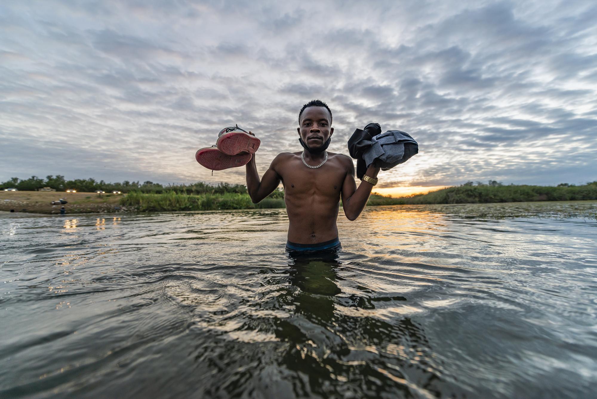 $!Migrante posa al amanecer, sujetando sus pertenencias en la línea fronteriza que cruza el río.