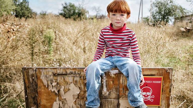 Un niño de la región de Kharkiv, Ucrania, sentado frente a un cartel que dice “Peligro de minas”.